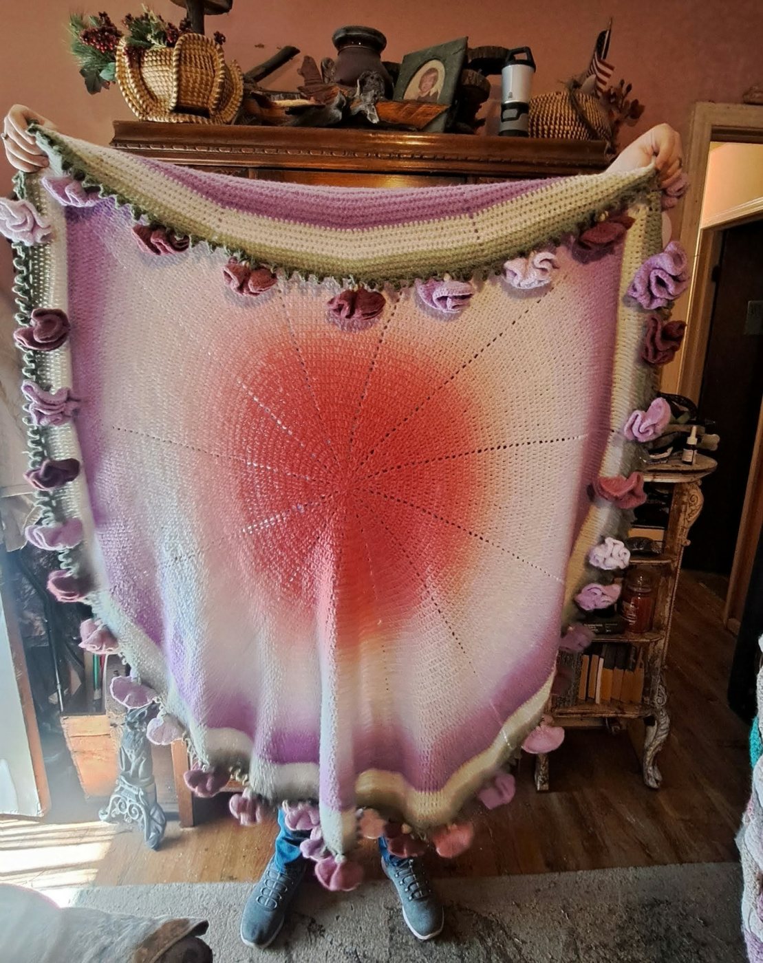 A round crochet blanket held open: pink at the centre fading to lilac and white, with a border of crochet roses