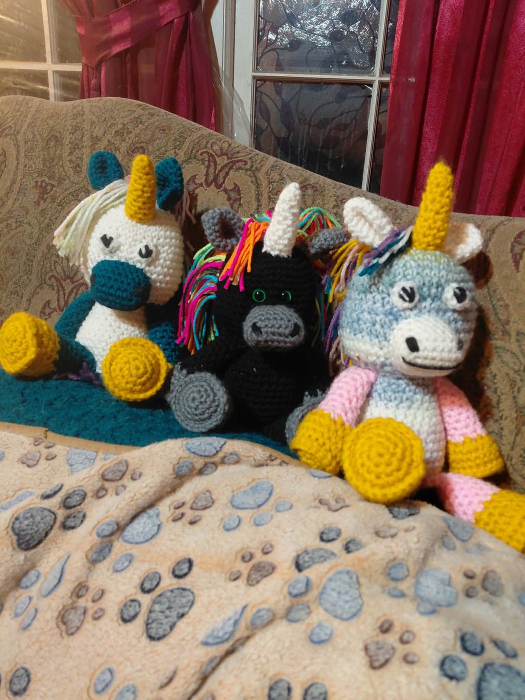 Three crochet unicorns on a sofa: white and teal, black with a rainbow mane, grey and pink